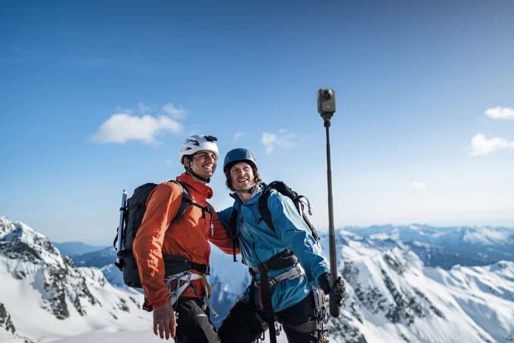 Dois homens a praticar montanhismo, a segurar um selfie stick com a câmara DJI Osmo 360 montada, com uma paisagem de montanhas e neve ao fundo.